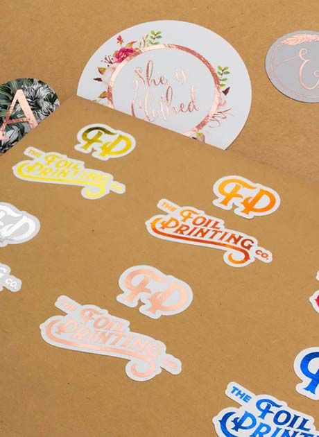 Collection of custom shaped metallic foil stickers displayed on a kraft background, showcasing branding, packaging, and promotional sticker applications.