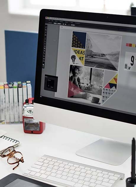 Computer screen displaying a print design layout on a desk with keyboard, glasses and stationery, representing preparing artwork files for print