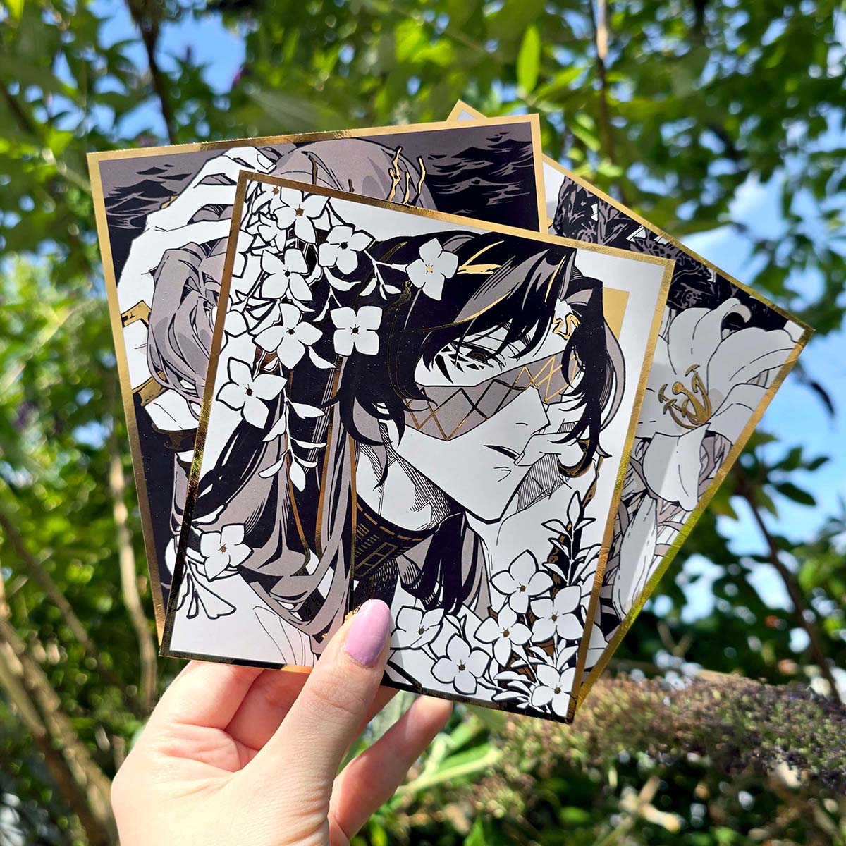 Metallic gold square anime art prints with monochrome character illustrations, floral details, gold foil borders and decorative linework.